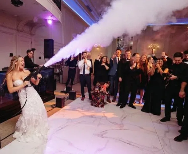 Een bruid in bruidsjurk vuurt Co2 rookkanon af op dansvloer terwijl gasten juichen. Op de achtergrond draait een bruiloft dj achter de dj booth.